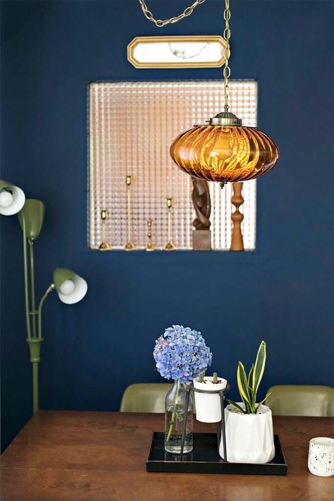 A dark blue wall with a vintage mirror hanging on it and a vintage lamp hanging in front.
