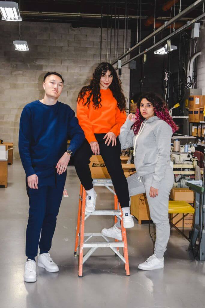Two women and one man standing in a warehouse wearing sweatshirts and sweatpants.