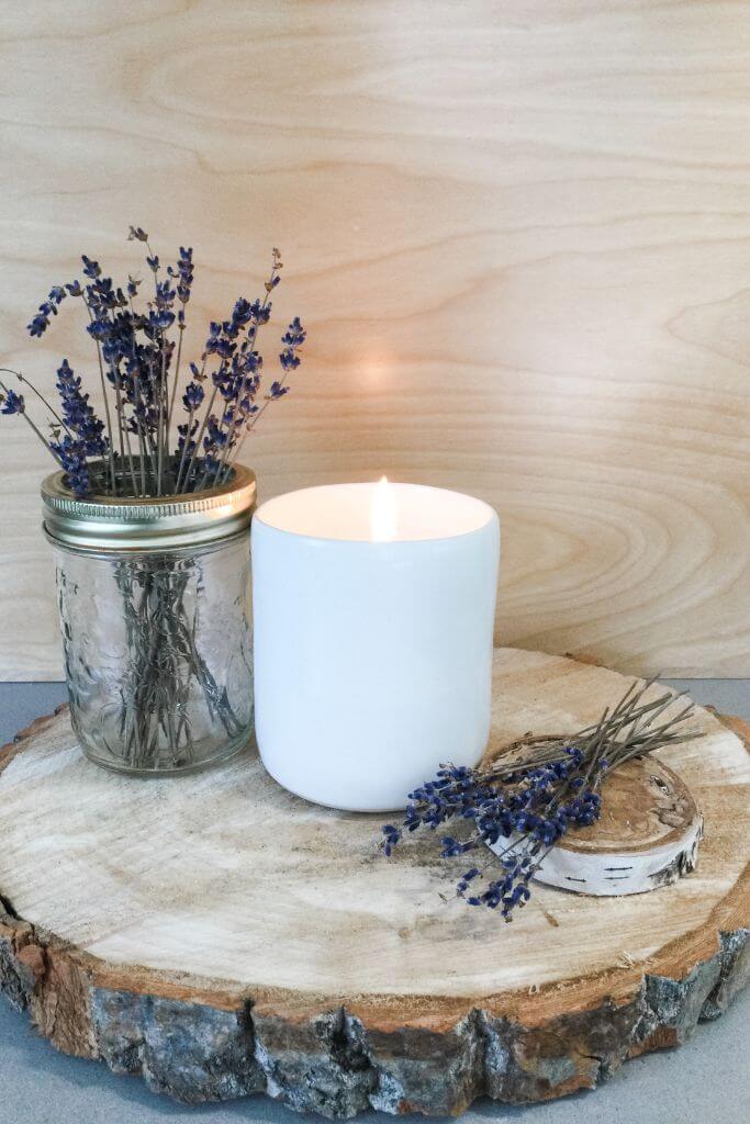 A candle in a white vessel sitting next to dried lavender on top of a wood slice.