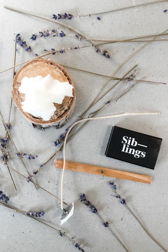 Candle wax, a cotton wick a wooden stick and a package of matches laying next to dried lavender.