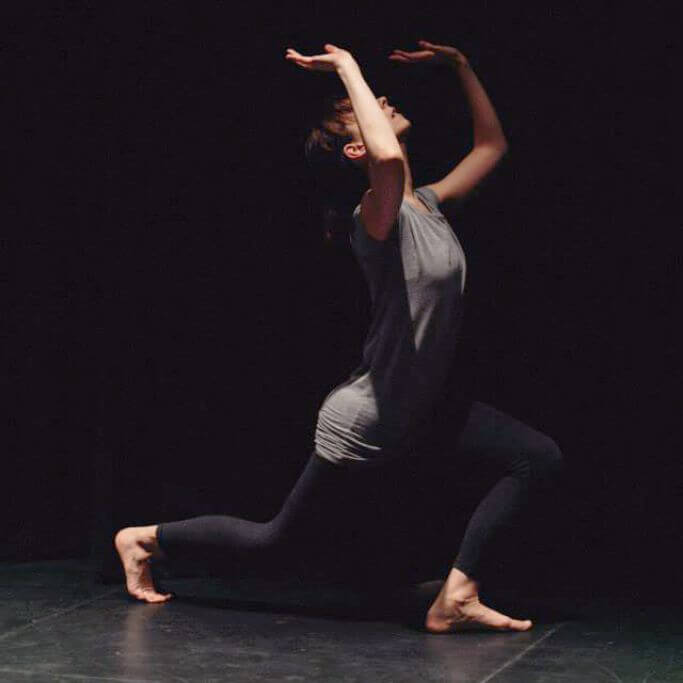 A dancer wearing grey clothing on a stage with a black background.
