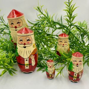 Five different-sized wooden Santa dolls next to greenery.
