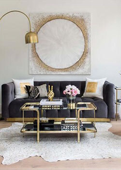 A glam-inspired living room with a grey couch, a glass and gold coffee table and a glittery painting.