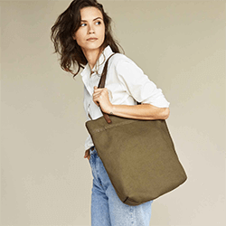 A woman carrying a green canvas bag wearing a white button-up shirt and jeans. Click to visit Nisolo website.