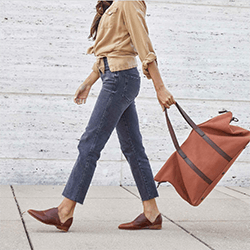 A woman carrying a brown canvas bag wearing loafers, jeans and a beige jacket. Click to visit Nisolo website.