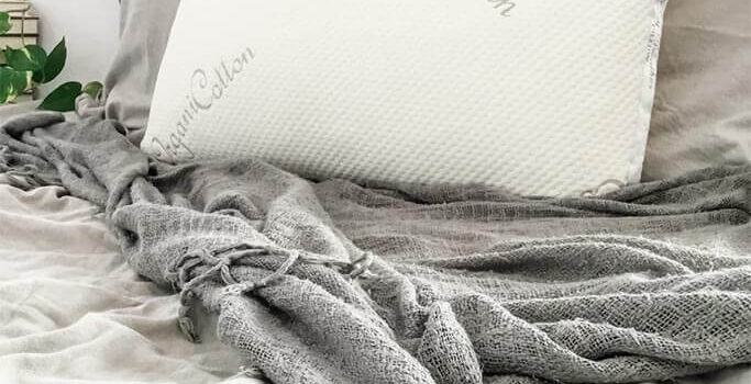 A white pillow sitting on a bed with grey sheets and blankets.