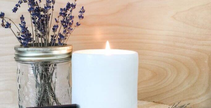 A candle in a white vessel sitting next to dried lavender on top of a wood slice.