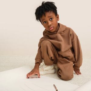 A little boy wearing a brown sweatsuit kneeling next to a paper and pencil.