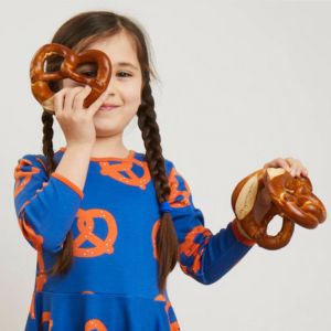 A little girl wearing a blue dress holding large soft pretzels.