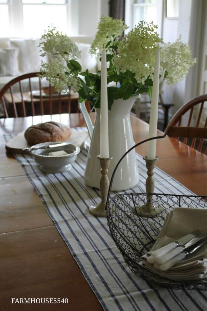 A wooden dining table with a striped blue and white runner, fresh flowers in a vase and white candles in brass holders.