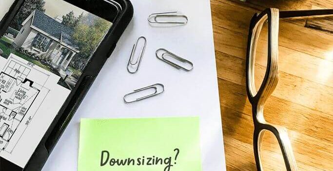 Office supplies such as paperclips and a pen sitting on a wood table with a plant, a pair of glasses and a tape measurer. A green note with the word "downsizing?" sits next to a phone with a floor plan on the screen.