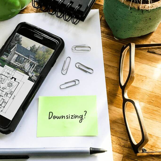 Office supplies such as paperclips and a pen sitting on a wood table with a plant, a pair of glasses and a tape measurer. A green note with the word "downsizing?" sits next to a phone with a floor plan on the screen. 
