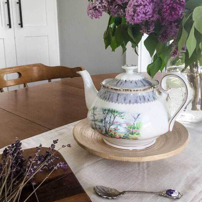A wooden dining table set with a cream coloured runner, fresh lilacs in a mason jar, a vintage teapot and dried lavender.