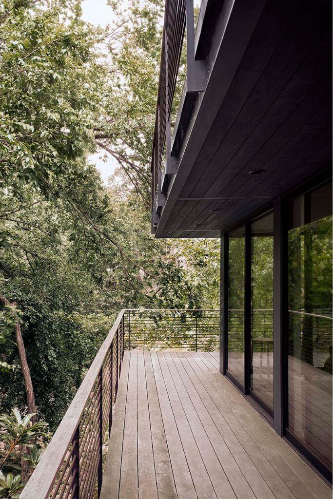 A light wood deck wrapping around a home with black-framed windows, surrounded by trees.