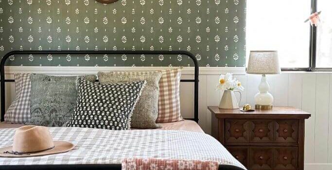 A bedroom with green floral wallpaper, a black metal bed and a mixture of patterned bedding.