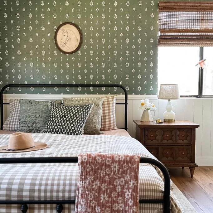 A bedroom with green floral wallpaper, a black metal bed and a mixture of patterned bedding.