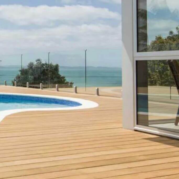 A light coloured wood deck next to a pool and a house with glass walls.