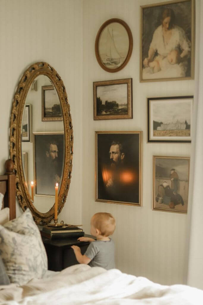 A little boy in a bedroom with lightly striped wallpaper and vintage art on the walls.