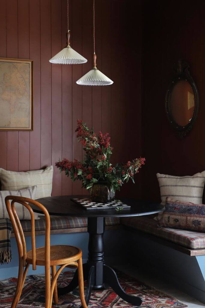 A dining area with dark purple walls, a small black table and a bench covered with vintage throw pillows.