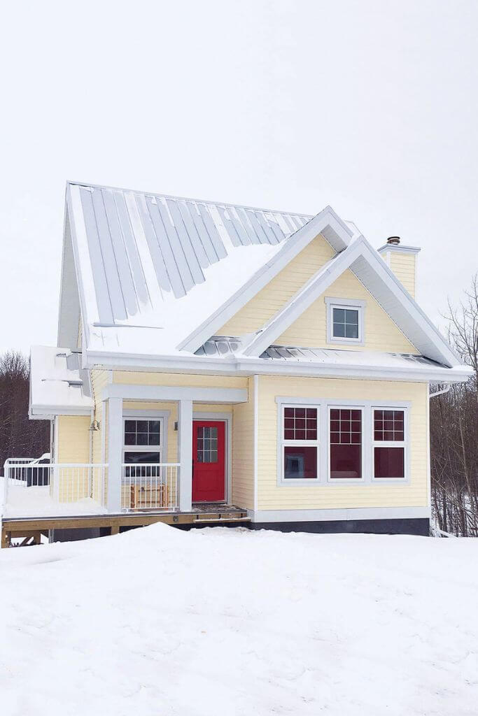 A yellow house with a metal room and red door in the winter.