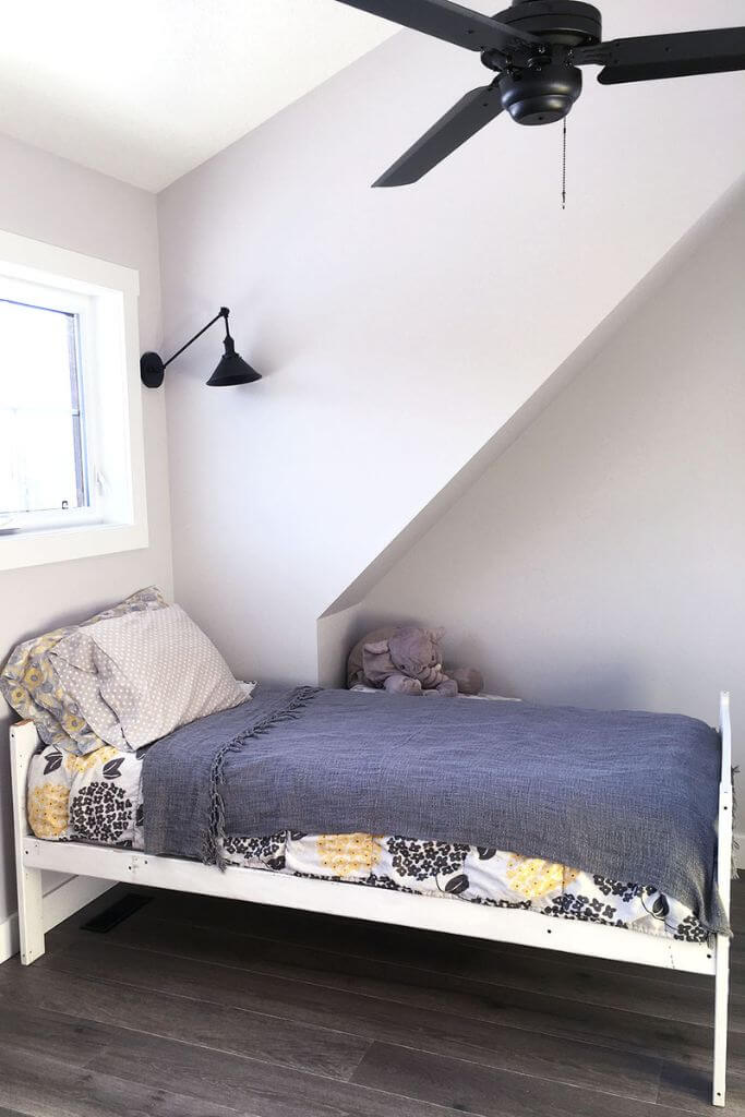 A room with a small white bed with grey, white and yellow bedding.