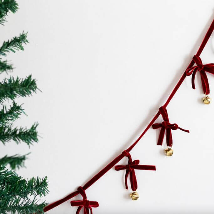 A red velvet and brass bell garland hanging on a wall near a Christmas tree.