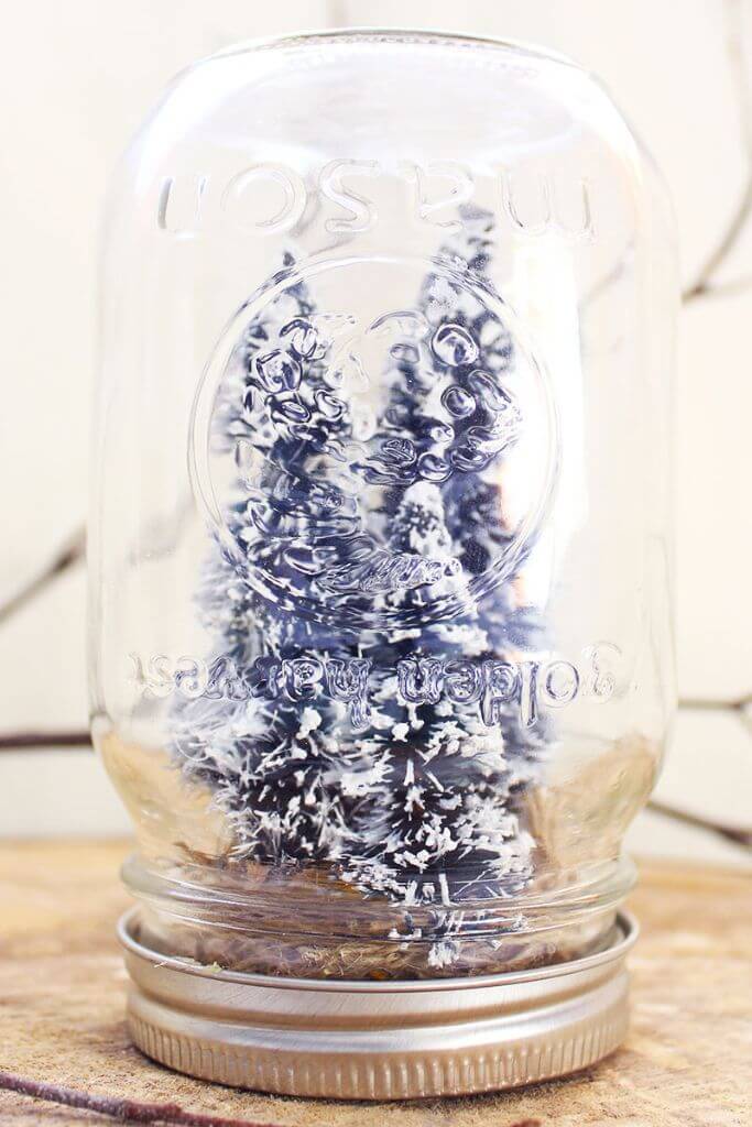 A handmade mason jar snow globe sitting on top of a tree slice in front of a twig.