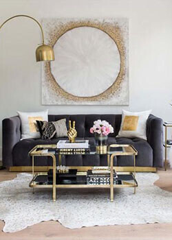 Image Credit: HF Home A glam-inspired living room with a grey couch, a glass and gold coffee table and a glittery painting.