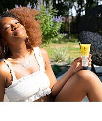 A young woman smiling in the sun holding a bottle of sunscreen. Click to visit the Beauty by Earth website.