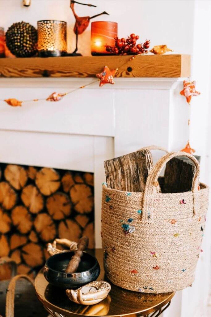 A woven storage basket with colourful flecks holding chopped wood next to a fireplace.